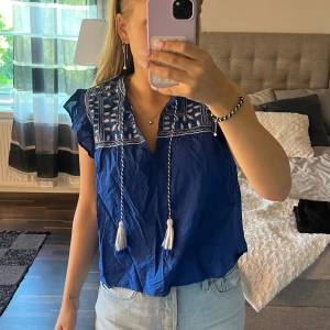Blå broderad blus från H&M - Säljer en blå blus från H&M med vit broderad detalj upptill och snören med tofsar. Blusen har korta volangärmar och v-ringad hals. Knappt använd 💙