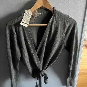 Grå omlottkofta med knyt i midjan - Snygg grå omlottkofta från Cubus med v-ringad hals och knyt i midjan. Koftan har långärmade ärmar. Har aldrig använt då jag ba ansåg att den inte helt passade mig. 