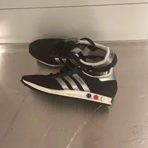 Adidas LA Trainer sneakers blå/silver - Säljer ett par Adidas LA Trainer sneakers i mörkblå mocka med silvriga detaljer och klassiska tre ränder på sidan. Skorna har vit sula med färgglada pluggar i blått, rött och vitt. Perfekt för dig som gillar retrostil och sportig vibe.