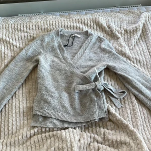 Grå omlottkofta från Gina Tricot - Säljer en mjuk och mysig grå omlottkofta från Gina Tricot i storlek S. Koftan har långa ärmar, ribbade muddar och knytband i sidan. Perfekt för lager-på-lager och chill dagar. Materialet är stickat och känns riktigt skönt mot huden.