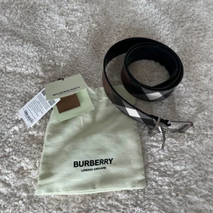 Burberry rutigt bälte i skinn - Snyggt Burberry-bälte i mörkbrunt och svart med klassiskt rutigt mönster. Bältet är gjort i skinn och canvas, har en silverfärgad metallspänne och kommer med originalpåse och taggar. Perfekt accessoar för att lyfta din outfit!