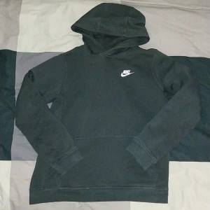 Svart Nike hoodie  - Svart hoodie från Nike i storlek 158-170, med klassisk vit Nike-logga på bröstet. Tröjan har huva och en stor magficka framtill. Tillverkad i mjuk bomullsmix, perfekt för chill dagar eller när du vill ha en enkel streetstyle-look.
