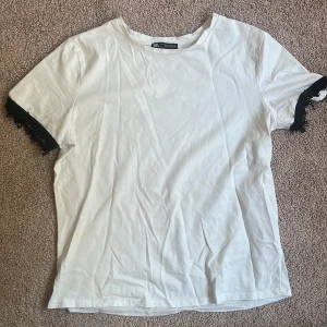 Vit t-shirt med spetsdetalj från Zara - Vit t-shirt från Zara i mjuk bomull med korta ärmar. Ärmsluten har svart spets som ger en cool kontrast och lite extra edge. Enkel rund halsringning och rak passform, perfekt till jeans eller kjol. Snygg och stilren med en twist.