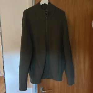 Olivgrön stickad kofta från Uniqlo XL - Säljer en olivgrön stickad kofta från Uniqlo i storlek XL. Koftan har ribbad struktur, hög krage och hel dragkedja framtill. Perfekt för lager-på-lager och chill stil. Materialet är mjukt och håller värmen, passar dig som gillar enkel och clean look.