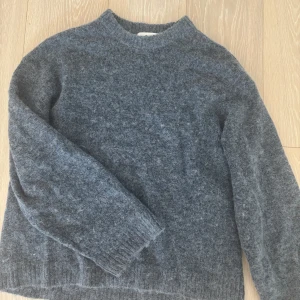 Grå/Blå stickad tröja från Gina Tricot - Mysig grå/blå stickad tröja från Gina Tricot i ullblandning. Tröjan har en enkel, rak passform med ribbade muddar vid hals, ärmslut och nederkant. Perfekt för kyliga dagar och enkel att matcha med jeans eller kjol.