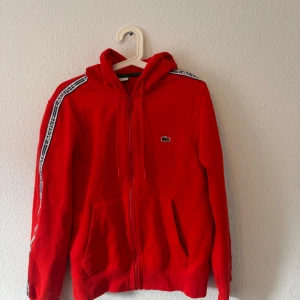 Röd hoodie från Lacoste med dragkedja - Säljer en röd hoodie från Lacoste med dragkedja framtill och snörning vid huvan. Tröjan har vita och svarta detaljer på axlarna med Lacoste-logga, samt klassisk krokodil på bröstet. Perfekt för chill dagar och streetstyle.