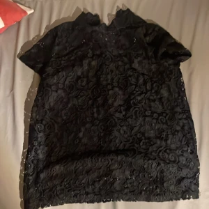 Svart spetsblus från STOCKH LM - Svart blus med korta ärmar från STOCKH LM, täckt av snygg spets med blommigt mönster. Blusen har en hög hals och en synlig dragkedja i guld baktill som ger en cool touch. Perfekt för dig som gillar detaljer och vill sticka ut.