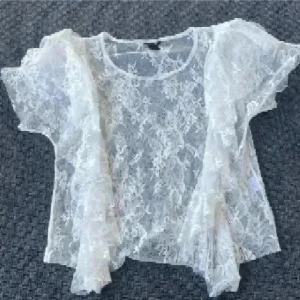 Vit spetsblus med volanger - Säljer en superfin vit blus i transparent spets med korta ärmar och snygga volangdetaljer längs sidorna. Blusen har rund halsringning och ett romantiskt mönster i spetsen som ger en extra cool vibe. Perfekt att styla lager på lager för en trendig look.