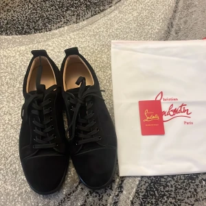 Svarta sneakers från Christian Louboutin - Säljer ett par svarta sneakers från Christian Louboutin med klassisk röd sula och snygg signatur på undersidan. Skorna har rund tå, snörning och är tillverkade i mocka med skinnfoder. Perfekt för dig som gillar exklusiv och stilren design.