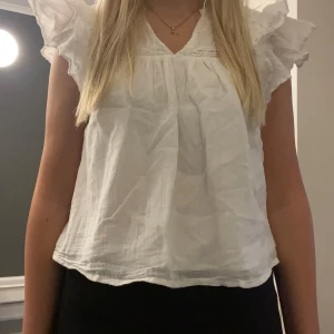 Vit blus med volangärmar - Supersöt vit blus med volangärmar och v-ringad hals. Blusen är lite croppad och har en luftig passform. Materialet känns som bomull och har en lätt skrynklig look. Perfekt för dig som gillar romantiska detaljer och vill ha en chill men ändå stylish topp.