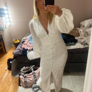 Vit långklänning med puffärmar - Säljer en vit långklänning med v-ringning, puffiga långärmar och knappar framtill. Klänningen har broderade blommor i tyget och en slits framtill. Passformen är normal och materialet känns som bomull. 