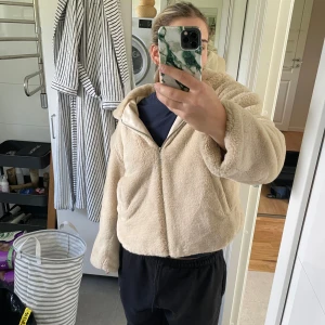 Beige teddyjacka med huva - Mysig beige teddyjacka med huva och dragkedja framtill. Jackan är croppad och har långa ärmar samt två fickor på sidorna. Perfekt för dig som gillar en chill och trendig look.