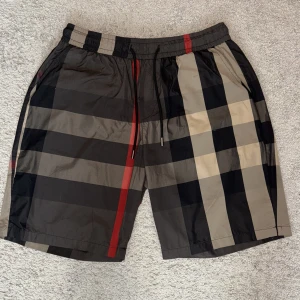 Blåa rutiga burberry shorts - Snygga Burberry shorts med klassiskt rutigt mönster i svart, beige och röd. De har elastisk midja med snörning och är tillverkade i ett lätt, glansigt syntetmaterial. Perfekt för dig som vill ha en clean och lyxig streetstil.