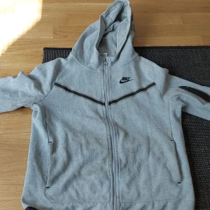 Grå Nike hoodie med dragkedja - Grå hoodie från Nike med svart logga på bröstet och svarta detaljer längs bröstet och ärmen. Tröjan har huva, hel dragkedja och två fickor fram. Materialet är mjuk bomull och polyester, perfekt för chill eller träning. Långärmad och med snygga svarta muddar.storlek 160