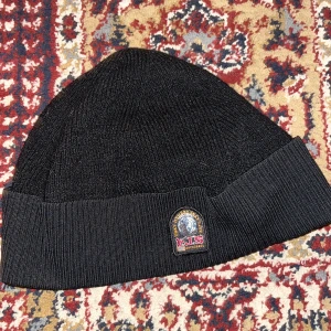 Svart mössa från Parajumpers - Svart ribbstickad mössa från Parajumpers med snyggt broderat märke framtill. Klassisk uppvikt kant och mjukt material som håller dig varm. Perfekt för dig som gillar stilrena accessoarer med en exklusiv touch.