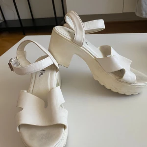 Vita sandaler med klack från Duffy - Säljer ett par vita sandaler från Duffy med breda remmar och justerbart spänne runt ankeln. Sandalerna har en chunky klack och plattformssula som ger extra höjd och cool vibe. Perfekt för dig som gillar stilrena och trendiga skor med lite edge.