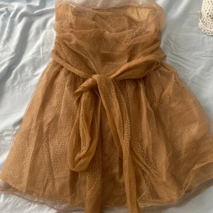 Champagnefärgad prickig kortklänning - Superfin champagnefärgad kortklänning med glittriga prickar och stor knytrosett i midjan. Klänningen är axelbandslös och har ett lager av transparent, skimrande tyg som ger en lyxig känsla. Perfekt för dig som vill sticka ut med en unik look.