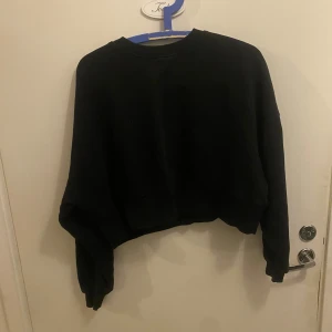 Svart croppad sweatshirt - Svart croppad sweatshirt med rund hals och långa ärmar. Tröjan har en loose passform och är tillverkad i mjukt bomullsmaterial, perfekt för chill dagar. Enkel och stilren design utan mönster eller detaljer, passar dig som gillar basic och comfy vibes.
