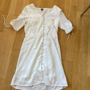 Vit kortklänning från Vero Moda XS - Supersöt vit klänning från Vero Moda i storlek XS. Den har korta ärmar med snörning, v-ringad hals och dekorativa klädda knappar framtill. Materialet är mjukt och har en lätt struktur som ger en lyxig känsla. Passformen är normal och klänningen slutar ovanför knäna.
