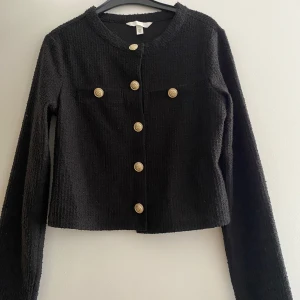 Svart cardigan jacka från H&M - Svart cardigan jacka från H&M med grovstickad struktur och snygga guldknappar framtill. Jackan har rund halsringning, lång ärm och två dekorativa fickor på bröstet💕köparen står för frakt! 