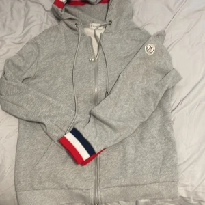 Grå Moncler hoodie med dragkedja - Snygg grå hoodie från Moncler med dragkedja och huva. Tröjan har coola detaljer som snörning vid halsen och Moncler-logga på ärmen. Muddarna har röd, vit och marinblå rand för extra stilpoäng. Perfekt för chill dagar eller när du vill se extra fresh ut.