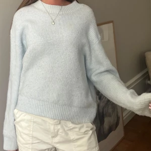 Ljusblå stickad tröja med rund hals - Mysig ljusblå stickad tröja med rundad hals och ribbade muddar vid ärmslut och nederkant. Tröjan har en loose passform och långa ärmar, perfekt för att chilla eller hänga med kompisar. Enkel och stilren design som är lätt att matcha med jeans eller shorts.
