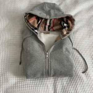 Grå Burberry hoodie med rutig huva - Säljer en grå hoodie från Burberry i storlek M. Tröjan har dragkedja, snörning i huvan och klassiskt Burberry-rutigt foder i huvan. Broderad logga på bröstet och två fickor framtill. Perfekt för dig som gillar stilren design med lyxig touch. Väldigt bra skick på hoodien och väldigt bra pris. Pris kan sänkas vid snabb affär!!!