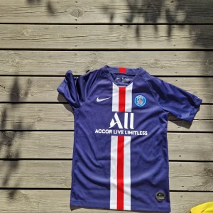 PSG Neymar 10 matchtröja Nike L - Snygg Paris Saint-Germain fotbollströja med Neymar 10 på ryggen. Marinblå med vit och röd vertikal rand på framsidan, PSG-emblem och Nike-logga. Tillverkad i lätt Dri-Fit material som andas, kortärmad och med klassisk rund hals. Perfekt för dig som älskar fotboll och PSG.