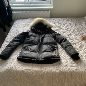 Svart dunjacka från Moose Knuckles - Säljer en svart dunjacka från Moose Knuckles med fluffig fuskpäls på huvan. Jackan har dragkedja, knappar och två stora fickor framtill. Insidan har coolt broderat märke och innerficka med dragkedja. Perfekt för kalla dagar och riktigt snygg streetstil.