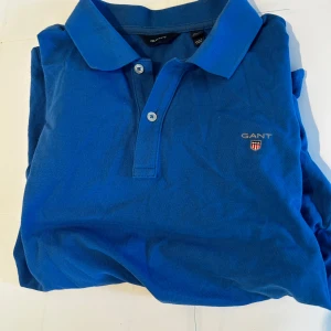 Blå pikétröja från GANT - Snygg blå pikétröja från GANT med klassisk krage och två knappar framtill. Tröjan har GANT-logga med broderat emblem på bröstet. Tillverkad i bomull som känns skön mot huden. Perfekt för dig som gillar stilren och prydlig look.