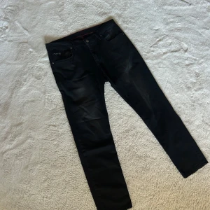 Svarta jeansbyxor från HUGO - Svarta jeansbyxor från HUGO med röd detaljsöm på insidan av midjan och broderad logga på bakfickan. Klassisk femficksmodell med raka ben och normal passform. Materialet är mjukt och stretchigt jeans, perfekt för en snygg och avslappnad look.