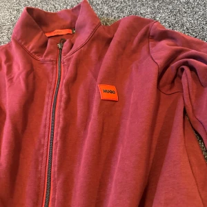 Röd zip-jacka från Hugo - Snygg röd zip-jacka från Hugo med stilren design och diskret logga på bröstet. Jackan har hel dragkedja framtill, två fickor med dragkedja och en hög krage. Perfekt för dig som gillar enkel och modern stil.