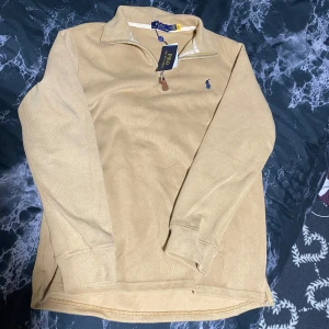 Beige half zip tröja från Polo Ralph Lauren - Snygg beige långärmad tröja från Polo Ralph Lauren med half zip och klassisk blå broderad logga på bröstet. Tröjan är gjord i mjuk bomull och har en stilren, normal passform. Perfekt för dig som gillar clean och enkel stil. Skriv gärna om mer bilder eller frågor.