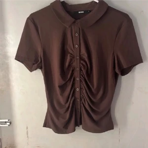 Brun blus med knappar från Bik Bok - Snygg brun blus från Bik Bok med korta ärmar och klassisk krage. Framsidan har cool rynkning och knappar hela vägen ner, vilket ger en unik look. Perfekt för dig som gillar detaljer och vill sticka ut lite extra. Materialet känns mjukt och skönt mot huden.