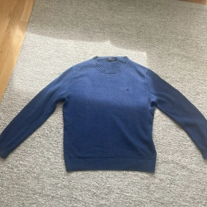Blå stickad tröja från Polo Ralph Lauren - Säljer en klassisk blå stickad tröja från Polo Ralph Lauren med rund halsringning och diskret broderad logga på bröstet. Tröjan har långa ärmar och ribbade muddar vid ärmslut och nederkant. Perfekt för lager-på-lager-stil. Storlek är M.