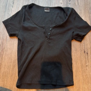 Svart ribbad topp från Gina Tricot - Svart ribbad topp från Gina Tricot i storlek S. Modellen är croppad med korta ärmar och har en v-ringad hals med tre knappar framtill. Materialet är mjuk bomull som sitter skönt mot kroppen och ger en slim passform. Perfekt för dig som gillar enkel och stilren vibe.