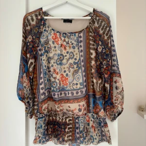 Blommig blus från Vero Moda - Säljer en luftig blus från Vero Moda i storlek L med färgstarkt blommigt och paisley-inspirerat mönster i blått, rött, beige och brunt. Blusen har trekvartsärmar, rund hals och volangdetalj nedtill. Materialet är tunn polyester som ger en skön och flowy känsla.