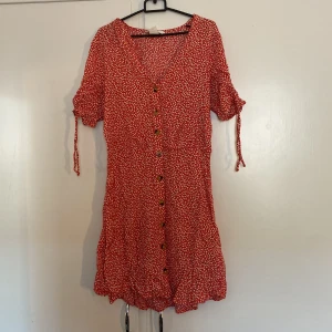 Röd blommig kortklänning från H&M - Superfin röd kortklänning från H&M med små vita blommor, v-ringning och knappar framtill. Klänningen har korta ärmar med snörning och en lätt volang nedtill. Perfekt för sommarens alla tillfällen!