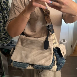 Beige och jeans axelväska med text - Unik axelväska/crossbody med beige flätad framsida och detaljer i blått jeansmaterial. Väskan har flera fickor, bruna och svarta detaljer samt bred axelrem i brunt tyg. Insidan är ljus och har dragkedja. Cool textprint på canvasdelen ger en edgy vibe. Ska se ”sliten ut”. 