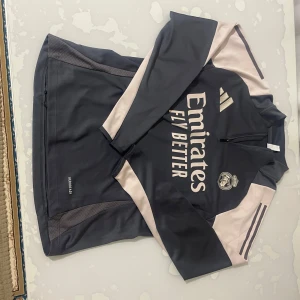 Adidas Real Madrid träningshoodie S - Snygg mörkblå och beige träningshoodie från Adidas med Real Madrid-logga och Emirates Fly Better-tryck på bröstet. Half zip framtill och långa ärmar med klassiska Adidas-ränder på axlarna. Perfekt för fotbollsträning eller chill. Materialet är polyester.