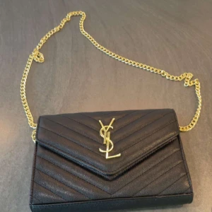 Ysl väska - Supersnygg svart kuvertväska från YSL med guldfärgad kedja och ikonisk logga framtill. Väskan har quiltad design och är tillverkad i skinn. Perfekt storlek för det viktigaste och ger en lyxig vibe till din outfit.