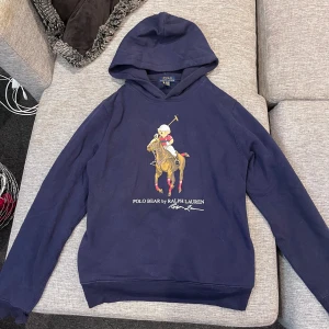 Mörkblå hoodie Polo Bear Ralph Lauren - Säljer en mörkblå hoodie från Polo Ralph Lauren. Framsidan har ett stort tryck med Polo Bear som spelar polo, och texten 'POLO BEAR by RALPH LAUREN'. Tröjan har huva och långa ärmar, tillverkad i mjuk bomull. Perfekt för dig som gillar klassisk streetstyle.
