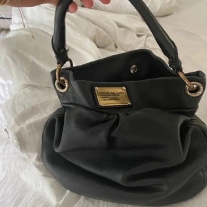 Väska Marc Jacobs - Cool svart handväska i mjukt skinn med guldfärgade metalldetaljer och en snygg logoplatta framtill. Insidan har ett mönstrat foder med stora bokstäver. Kan gå ner i pris vid snabb affär 