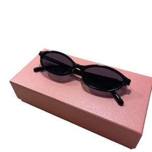 Svarta ovala solglasögon från Miu Miu - Snygga svarta solglasögon med smal, oval båge i blank plast. Glasen är mörka och ger en cool, retro vibe. Perfekta för dig som vill sticka ut med en trendig accessoar från ett lyxigt märke. Boxen på bilden medföljer ej! 