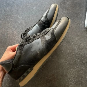 Valentino Rockrunner - Svarta Valentino Rockrunner sneakers med coola camouflagedetaljer och klassiska nitar vid hälen. Skorna har snörning, platt sula och är tillverkade i en mix av skinn och mocka. Snygg kontrast mellan svart ovandel och beige sula. Perfekta för dig som gillar streetstyle.