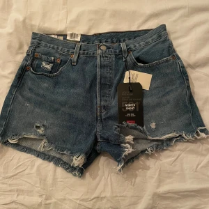 Levi's blå jeansshorts - Snygga blå jeansshorts från Levi's med råa, fransiga kanter och slitna detaljer. Klassisk femficksmodell med hög midja och knappgylf. Perfekta för en avslappnad och trendig look i sommar. Ikonisk Levi's-lapp bak. Hör av dig vid frågor 😁