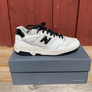 Vita New Balance sneakers  - Snygga vita New Balance sneakers med svarta detaljer och svart N-logga på sidan. Skorna har svarta snören, robust svart sula och är tillverkade i skinn och syntet. Perfekta för dig som gillar klassisk streetstyle med en sportig touch.