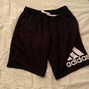 Svarta Adidas shorts - Svarta shorts från Adidas med vit snörning i midjan och stort vitt Adidas-tryck på ena benet. Sköna och sportiga, perfekta för träning eller att bara chilla i. Tillverkade i mjuk bomull för maximal komfort.