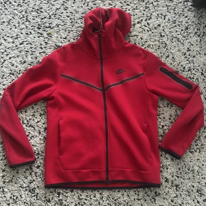 Röd Nike tech   - Nike vindjacka i rött med svarta detaljer och hel dragkedja. Jackan har huva, två fickor framtill och en cool svart rand över bröstet. Perfekt för dig som gillar sportig stil och vill sticka ut. Materialet är mjukt och känns skönt mot huden.
