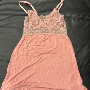 Rosa natt klänning  - Söt kort rosa natt klänning med smala axelband och vacker spets upptill. Klänningen har en mjuk, flowy kjol och detaljerad spets över bysten och midjan. Perfekt för dig som gillar romantisk och feminin stil. Har nått litet hål men syns knappast 
